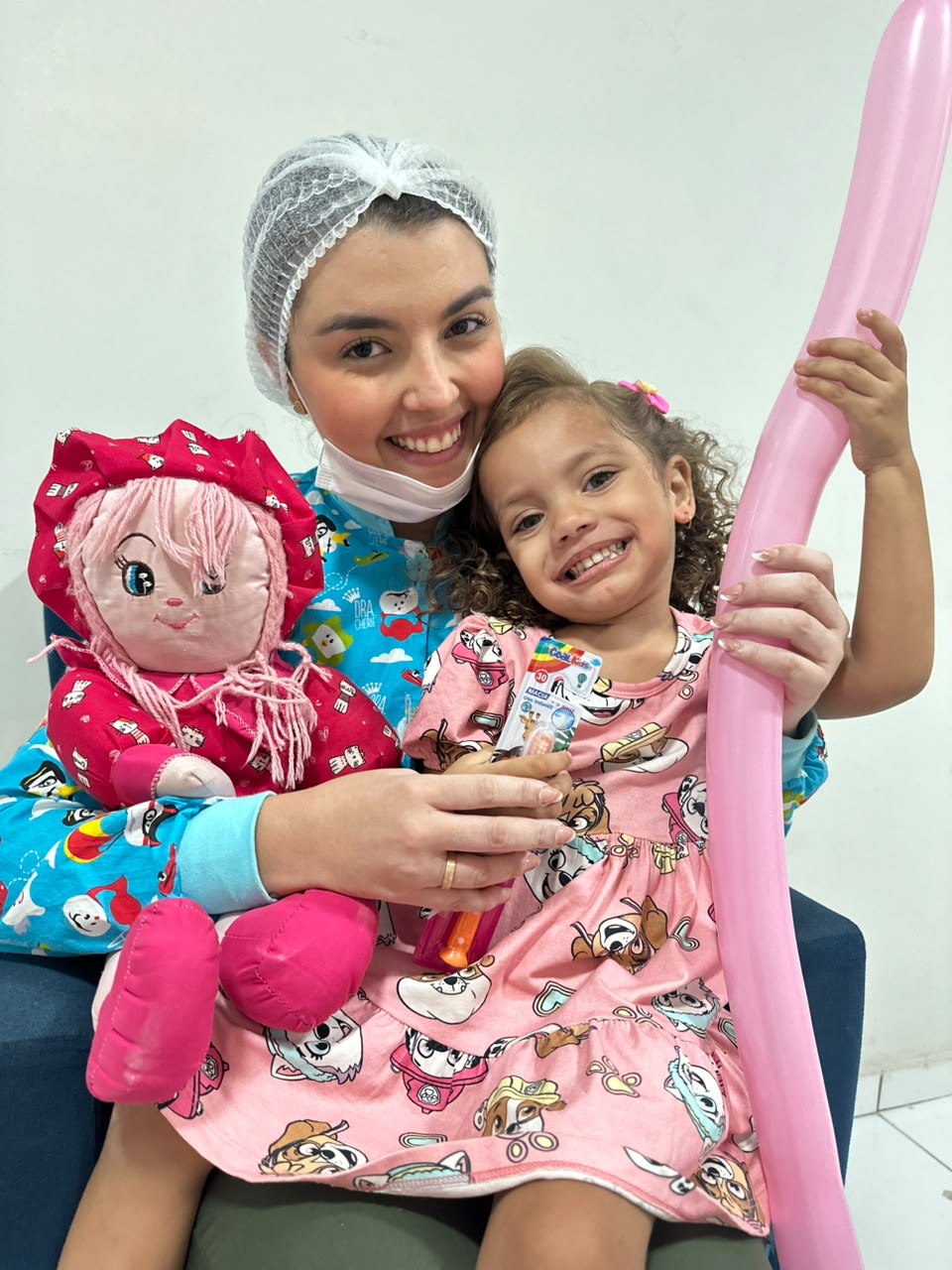 Dra. Thirza Nunes com uma menina sorrindo, boneca e balão na clínica infantil